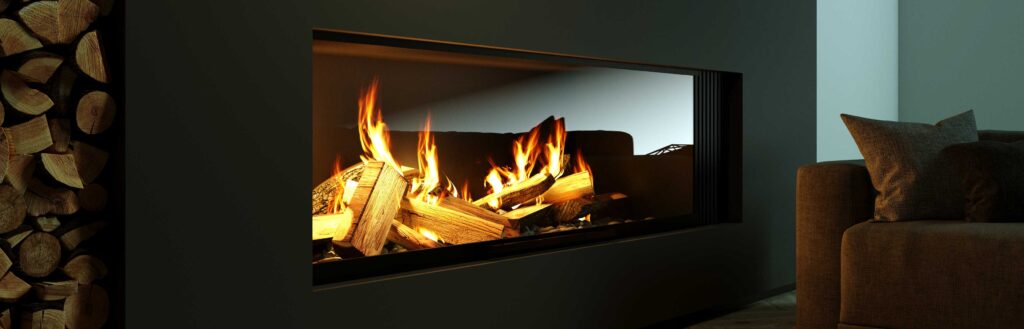 A modern fireplace with burning logs and bright flames, set in a dark wall. Firewood is stacked neatly on the left, and part of a gray sofa with cushions is visible on the right.