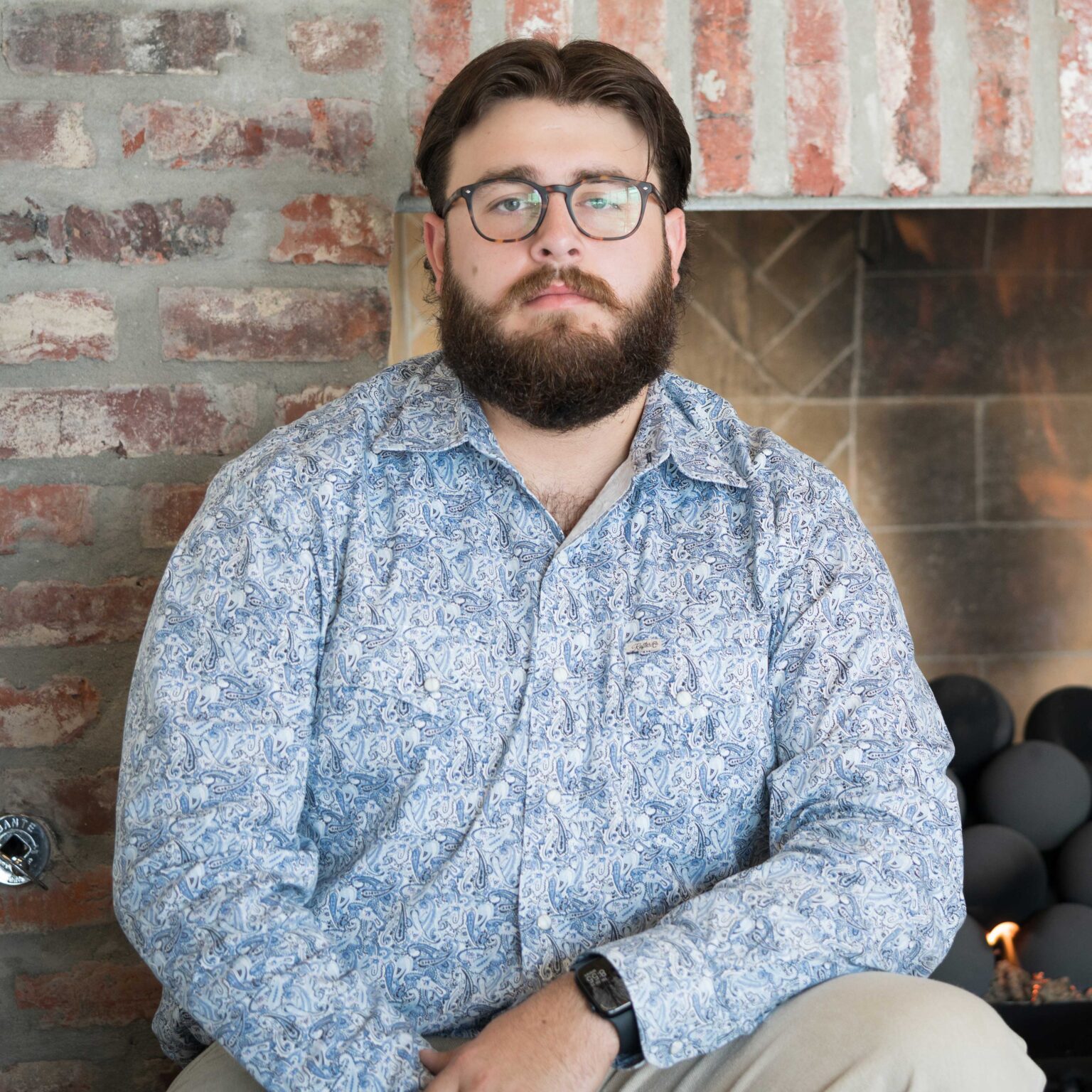 A man with a beard and glasses, wearing a patterned shirt and watch, sits in front of a brick fireplace with a neutral expression.